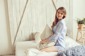 young woman in pajama wake up in the morning in cozy scandinavian bedroom and sitting on bed with white bedlinen. Casual lifestyle in modern interior