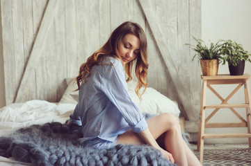 young woman in pajama wake up in the morning in cozy scandinavian bedroom and sitting on bed with houseplants on background. Casual lifestyle in modern interior