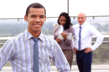 Holding a tablet, a young handsome black businessman is standing outside of a business building, confidently looking forward.