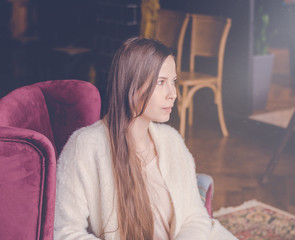 Fototapeta premium Beautiful girl dreaming about something while sitting in a cafe and looking at window. Young charming woman thinking about new ideas, drink a coffee, sitting on a lilac armchair in a modern city bar.