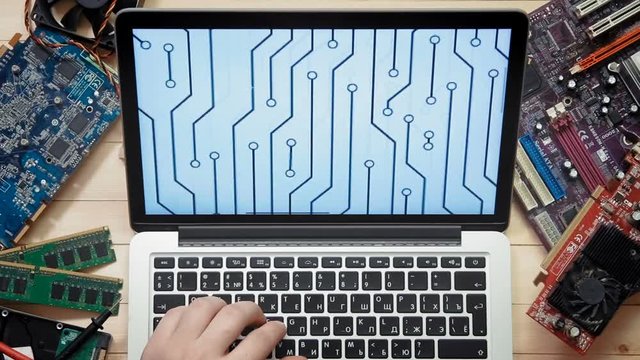Top view computer technician using laptop computer with circuit board outline at wooden desk with tools and electronic components