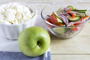 Foods for healthy eating: cottage chesse, apple, salad on light wood background