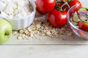 Foods for healthy eating: cottage chesse, tomatoes, apple, cereal, salad on light wood background