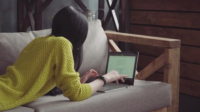 Young woman with long hair lies on the couch and works on the laptop, view from the back