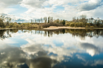 The bay of the river, in calm water reflects a blue sky with clouds, on the horizon there is a forest of deciduous and coniferous species, dry vegetation, a spring sunny day