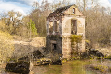 The old destroyed building of the hydroelectric plant with moss-grown walls is located in a forest surrounded by dry grass and shrubs, a sunny spring day, a blue sky with clouds