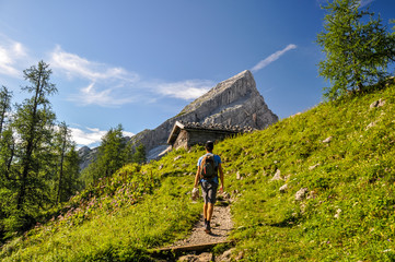 Young man hiking on a pathway up to Watzmann Mountain (2713m). In the background you can see a cabin and  "Kleiner Watzmann" (Little Watzmann, 2307m). Sunny day in the Bavarian Alps in Germany.