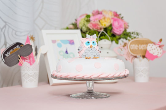 Birthday cake covered with fondant displayed on the pink cloth and glass tray; decorated with pink dots and an grey and pink fondant owl with blue eyes sitting on top