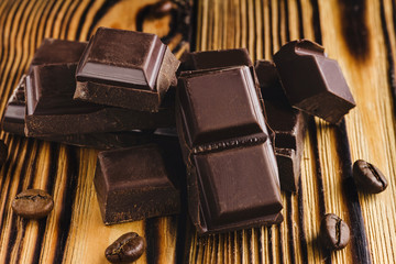 Pieces of bitter, dark chocolate, cubes on heap and coffee bean on wooden background, close-up view