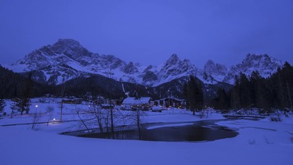 Day-to-night timelapse of wintry village at the foot of a snow-covered mountain.