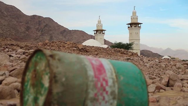 Iron barrel lies on the stony ground in defocus, mosque with two minarets on background in focus. Mosque and oil barrel. Old green rusty cask in mountain in Aqaba, Jordan. Empty barrel, lack of fuel
