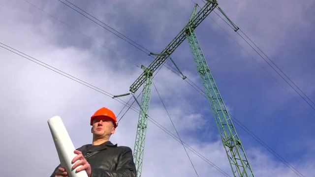 Electrical engineer in an orange helmet with a plan of electric grids in the hands under the power lines, low point shot