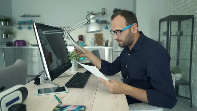 Unhappy, overwhelmed man working with documents and pc computer by table at home
