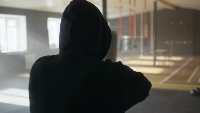 Slow motion shot with rear view of unrecognizable fighter in hoodie walking and warming up arms in dark gym lit by natural lighting coming from windows