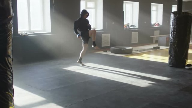 Slow motion of hooded boxer shadow fighting and warming up before practice in empty gym lit by natural lighting coming from windows