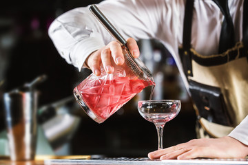 Male bartender is making cocktail pouring alchohol from jug to glass at bar background.