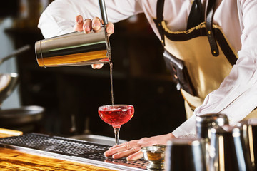 Male bartender is making cocktail pouring alchohol from shaker to glass at bar background.