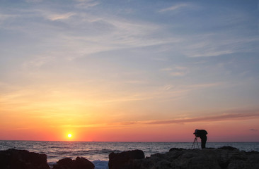 Silhouette of a photographer with a backpack and tripod with a beautiful sea sunset on the background