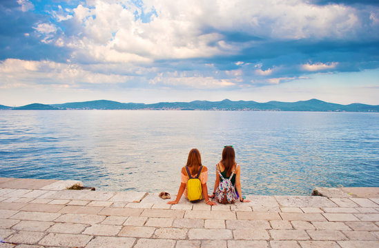 Two young girls with backpacks sitting closely to each other, their hands leaning against stone pavement, looking at the hill range in the distance. Back view. Town of Zadar, Croatia