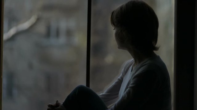 Depressed woman sitting on the sill and looking out the window, broken-hearted