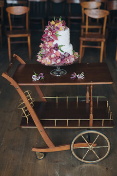 A two-level white wedding cake decorated with flowers and stands on wooden table on the background of vintage chairs in the wedding ceremony area