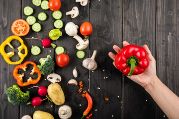 Naklejka na ściany i meble Top view of hand holding pepper and fresh vegetables on wooden table background