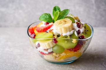 Fresh fruit salad with yogurt and walnuts in glass bowl on stone background.