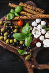 Mediterranean appetizer antipasti board with green black olives, feta cheese, mozzarella, capers, pepper, basil with grissini bread sticks over black wood burnt background. Top view with space