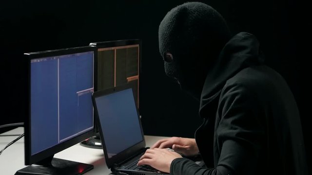 Hacker in balaclava cracking code using laptop and computers from his dark hacker room