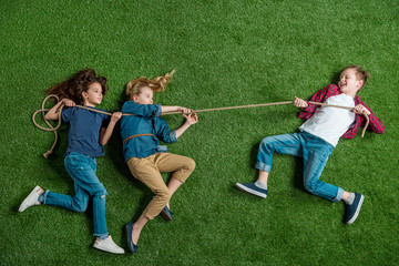 Top view of cute girls and boy lying on grass and playing tug of war