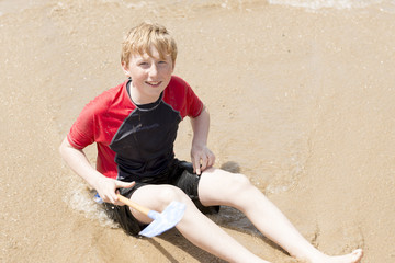 Cheerful Young Boy with Toy Shovel Sitting on Wet Sand