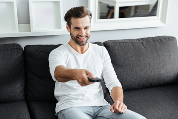 Fototapeta premium Young cheerful man sitting on sofa holding remote control.