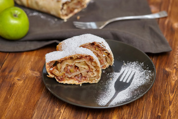 Apple strudel with icing sugar on black plate, wooden background