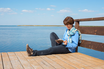 Dreaming woman is sitting on a wooden pier with tablet