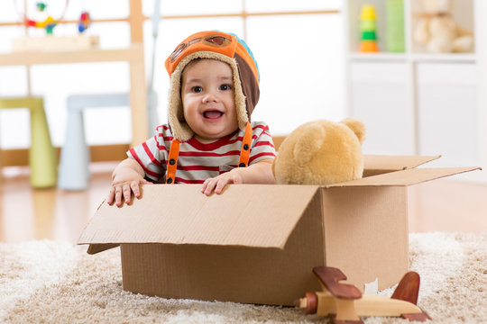 Smiling pilot aviator baby boy with teddy bear toy plays in cardboard box