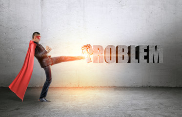 A businessman in a red cape and a mask beating a concrete 'Problem' writing with his leg.