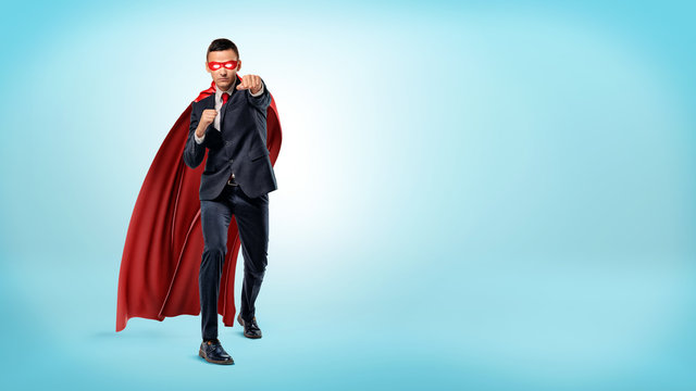 A businessman in a flowing red superhero cape and a mask throwing punches at an invisible enemy on blue background.