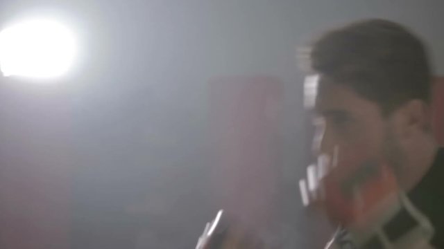 Giovane ragazzo si sta allenando al sacco da boxe in palestra con i guanti rossi e la maglia nera. Fumo bianco nello sfondo. 