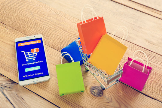 White smartphone runs an online shopping app put near colorful paper shopping bags and a shopping cart. Consumers can buy everything from online stores using a smart device connects to the internet.