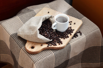 A full bag of brown coffee beans and a white cup of hot coffee lies on a wooden surface