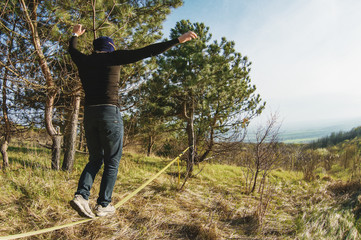 A man, aged with a beard and wearing sunglasses, balances on a slackline in the open air between two trees at sunset