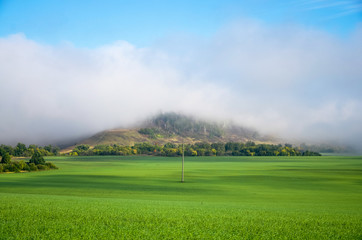 Green meadow under blue sky with a mountain in the middle and hanging over it a fog. Nature landscape.