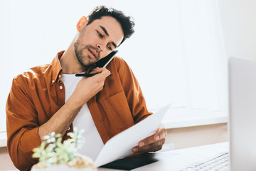 Fototapeta premium Freelancer communicate sales problems. Business, people, modern technology and communication concept. Attractive young entrepreneur having phone call, sitting in his office with notebook computer. 