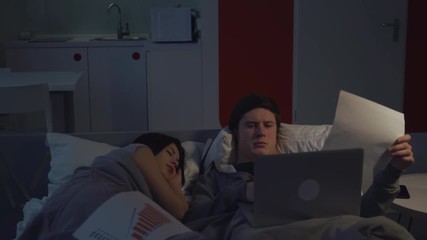 businessman working in bed. Young man using laptop lying near sleeping girlfriend.