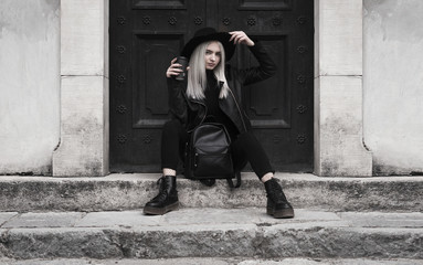 A stylish girl in a hat with white hair, dressed in black clothes, sits on the steps, holding a black coffee in her hand. Concept: only black.