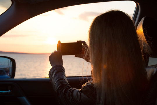 Young woman looking out car window of the sunset on the sea and taking pictures on your cell phone.