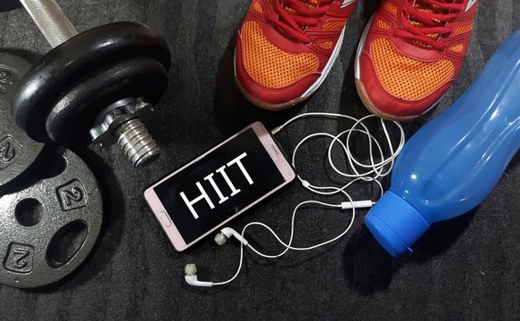 Healthy and fitness concept. Smartphone, dumbbell, water bottle and sport shoes on gym floor with word HIIT