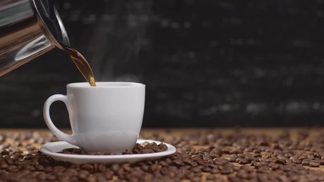 Pouring coffee from coffee pot in white cup surrounded by coffee beans on dark background with copyspace. 4k UHD