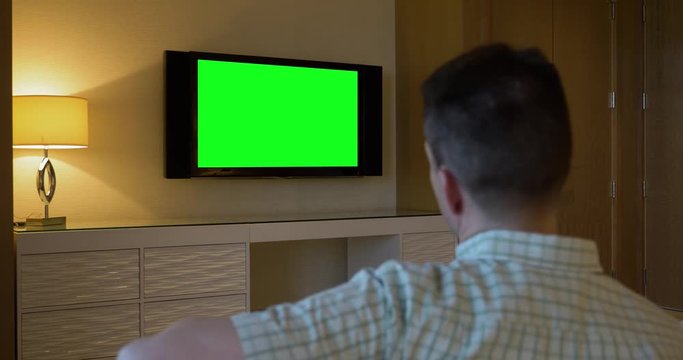 A man watches a television in a luxury hotel room. From behind, with green screen for customizable content.  	