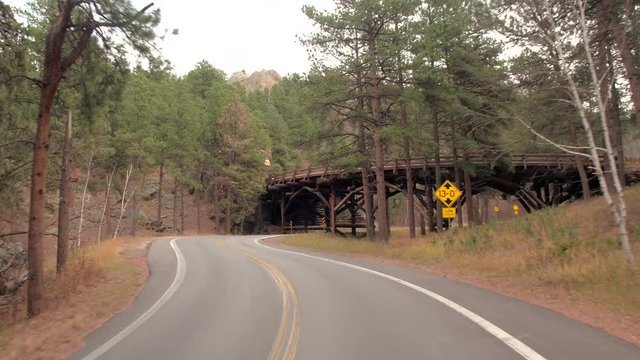 FPV, CLOSE UP: Driving under the wooden pigtail bridge on curvy serpentine Iron Mountain Highway surrounded by beautiful pine forest in Black Hills, South Dakota, USA. Detail of log pillars and arches